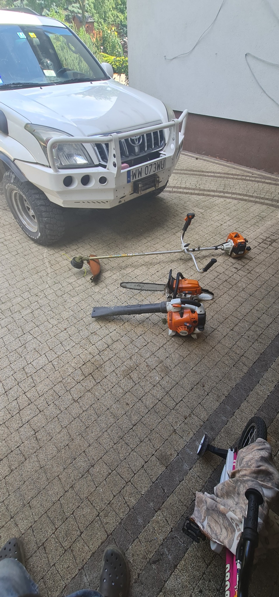 Na brukowanym podjeździe przed białym samochodem terenowym leżą: kosa spalinowa, dmuchawa do liści i piła łańcuchowa marki Stihl, obok zaparkowany rower dziecięcy.