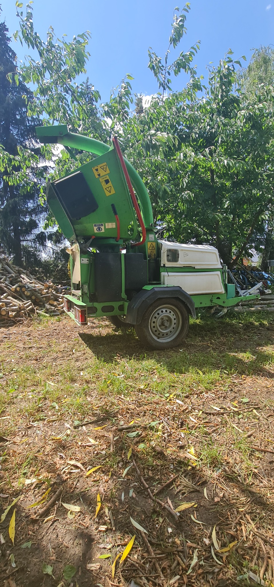 Zielono-biała rębarka do gałęzi na przyczepie, otoczona ściętymi gałęziami i drzewami w słoneczny dzień.