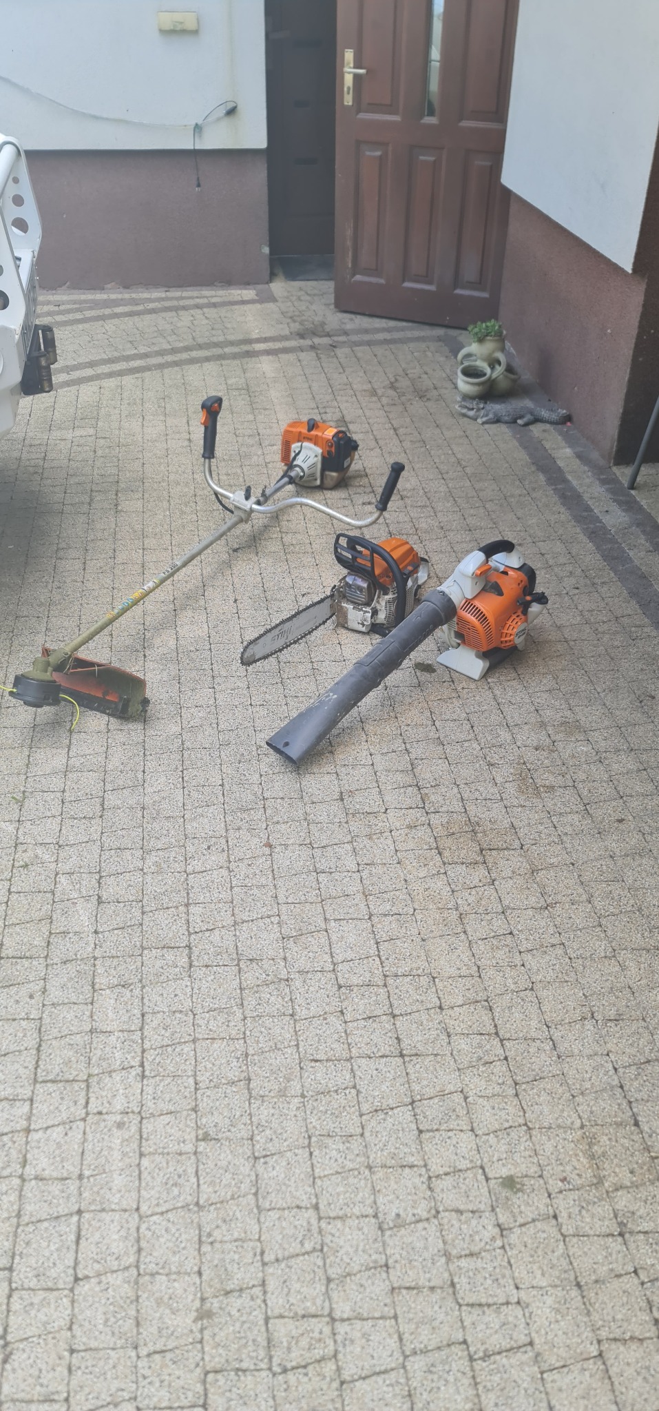 Różne narzędzia ogrodnicze Stihl leżące na kostce brukowej przed domem: kosa spalinowa, dmuchawa do liści i piła łańcuchowa.