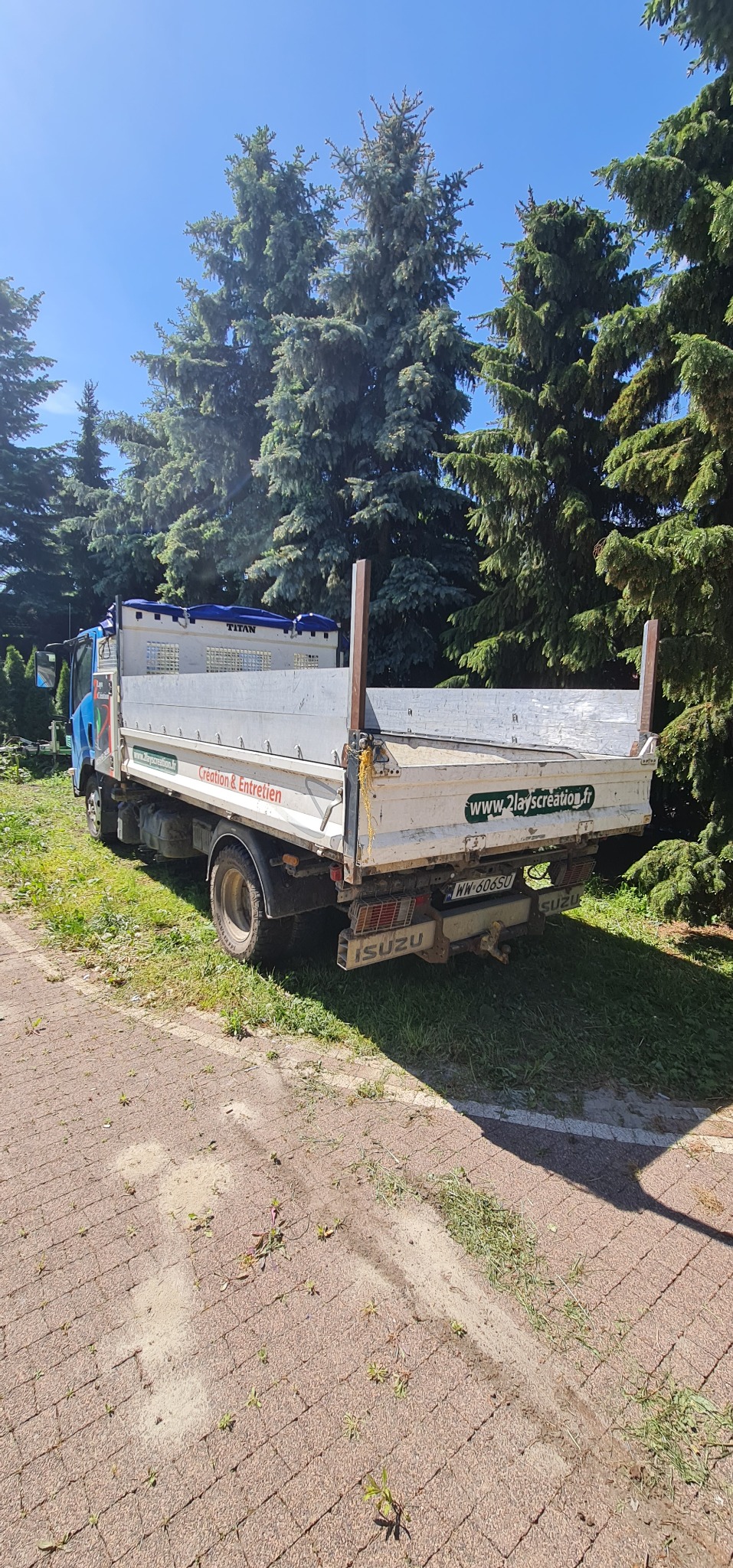 Isuzu z otwartą skrzynią ładunkową, zaparkowany na brukowanej ścieżce z widocznym logo firmy i adresem strony internetowej na klapie, w tle wysokie drzewa iglaste i błękitne niebo.