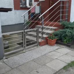 Wejście do domu z niską, nierdzewną furtką i balustradą schodową o minimalistycznym designie, z widocznymi detalami mocowania do ceglanej elewacji.