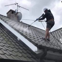 Pracownik w kasku i uprzęży spryskuje dach z szarej dachówki wodą pod ciśnieniem, widoczny komin z anteną i rynna.