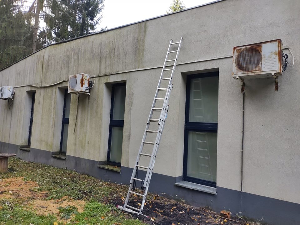 Zewnętrzna ściana budynku z widocznymi zabrudzeniami i rdzewiejącymi jednostkami klimatyzacji; aluminiowa drabina oparta o ścianę, sugerująca prace konserwacyjne.