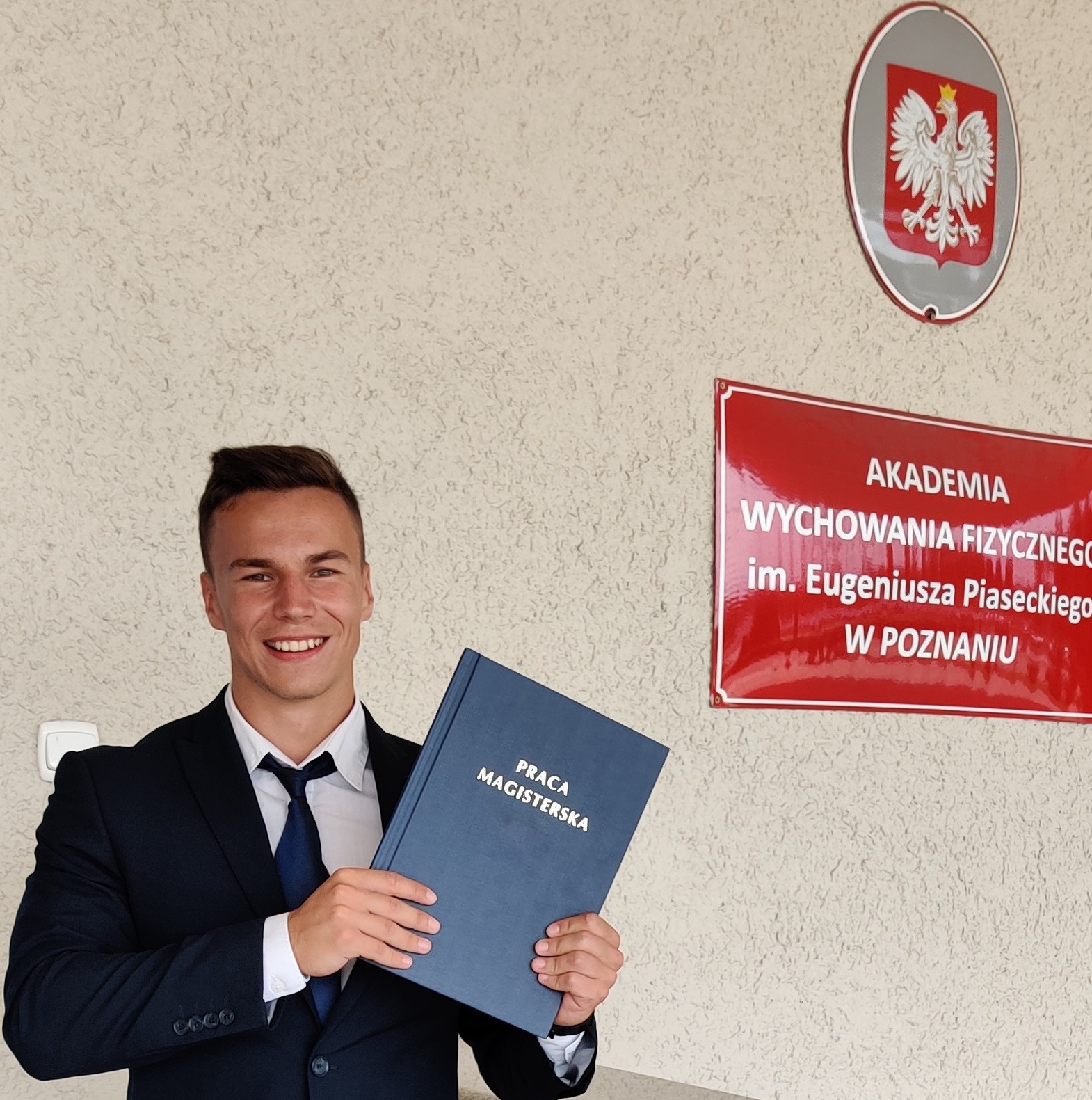 Uśmiechnięty absolwent w garniturze trzymający pracę magisterską przed budynkiem Akademii Wychowania Fizycznego w Poznaniu.