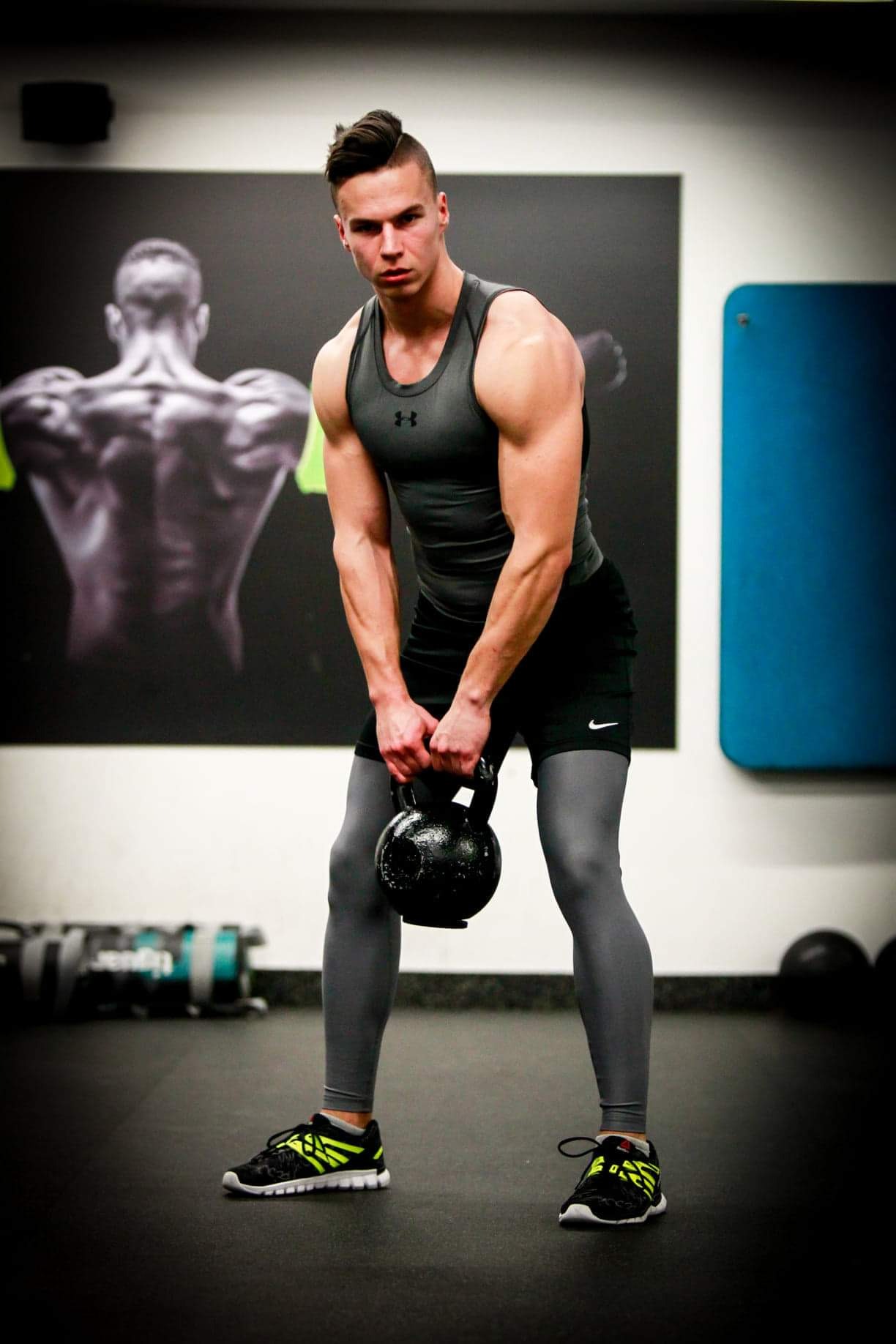 Mężczyzna w sportowym stroju, z napiętymi mięśniami, podnosi czarny kettlebell w siłowni; w tle plakat z sylwetką sportowca.
