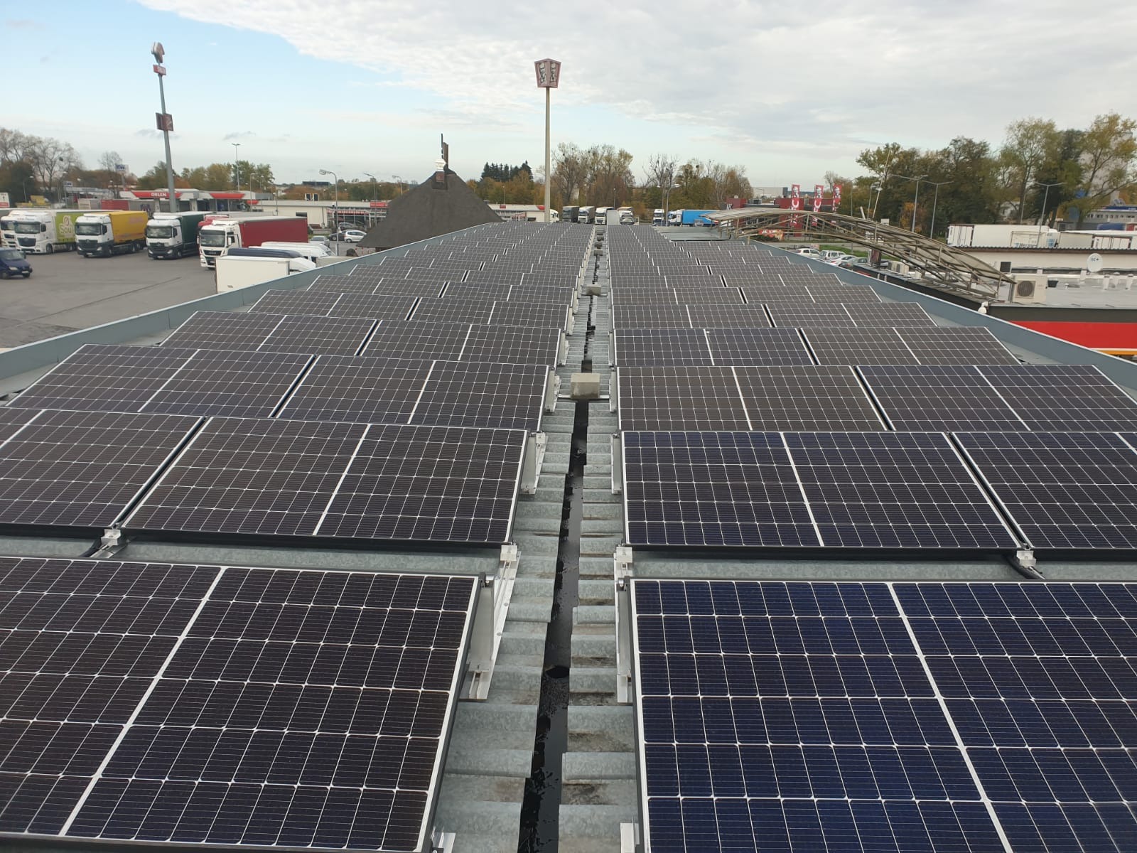 Rozległa instalacja paneli słonecznych na dachu budynku, widok z góry, w tle parking z samochodami ciężarowymi i budynki.