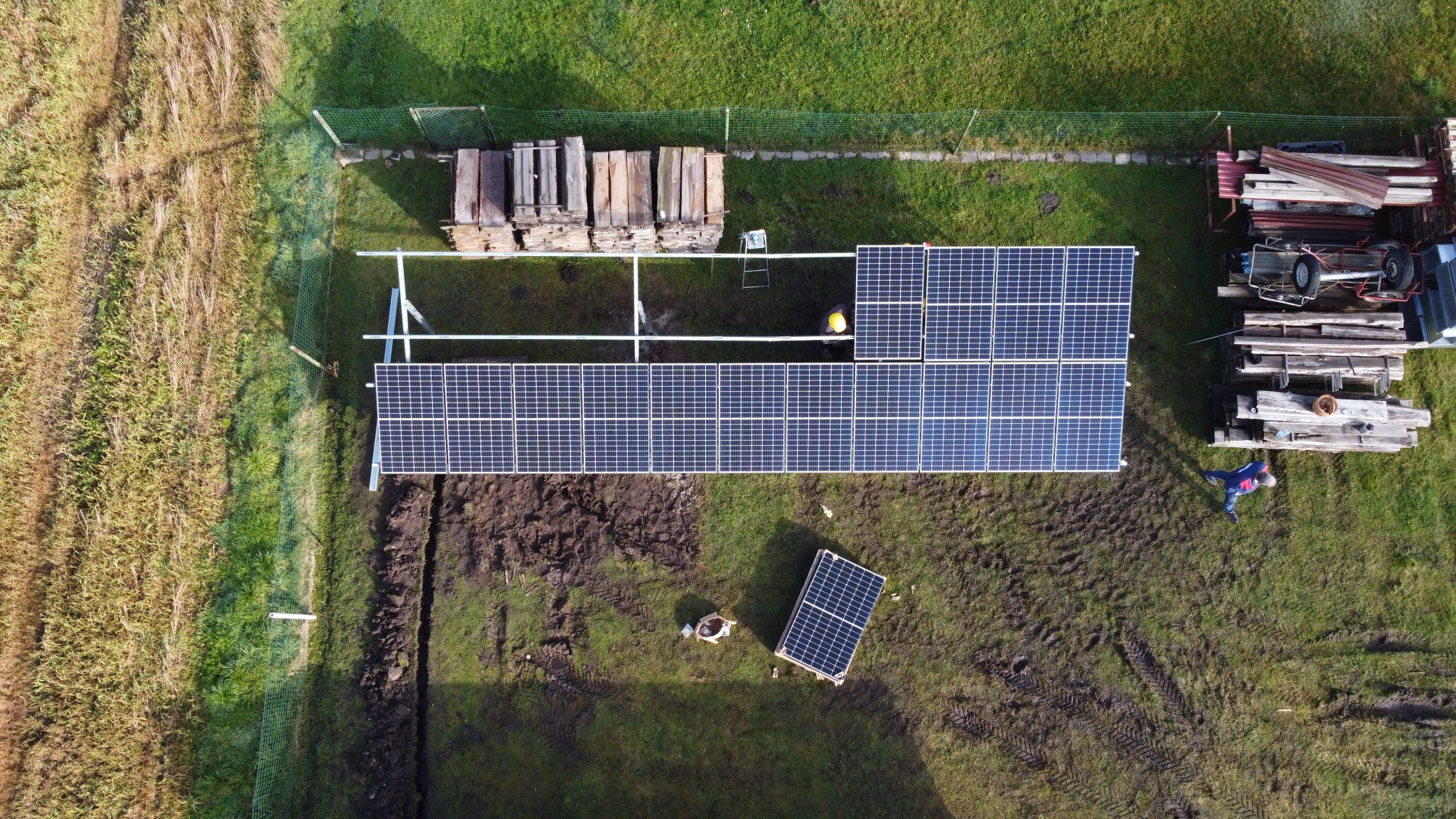 Widok z drona na montaż paneli słonecznych na trawiastym terenie, widoczna konstrukcja wsporcza, stosy drewna, pracownik w niebieskiej kurtce oraz pojedynczy panel na ziemi.