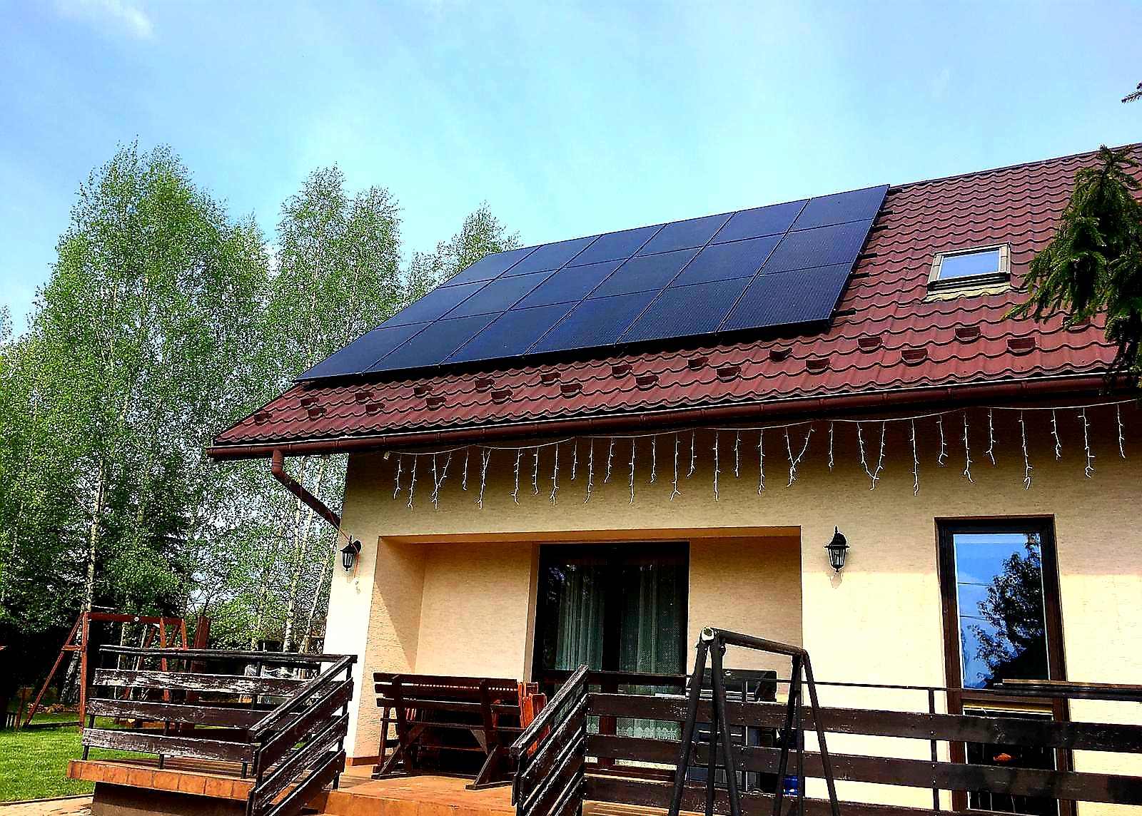 Panele słoneczne na dachu domu z czerwoną dachówką, widok z ogrodu z drewnianym tarasem i huśtawką.