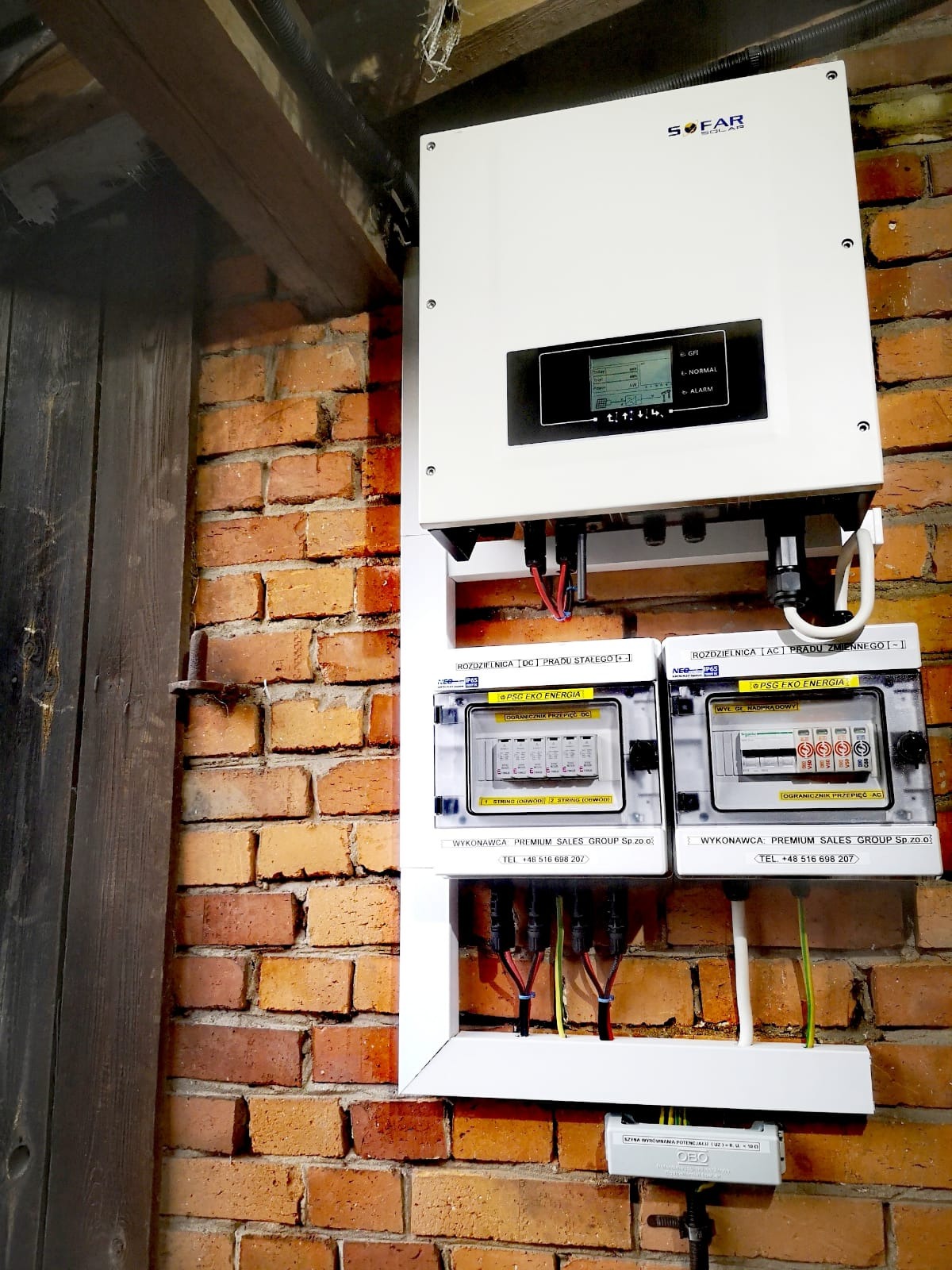 Biały falownik Sofar Solar i rozdzielnice prądu stałego i zmiennego na ceglanej ścianie wewnątrz budynku.