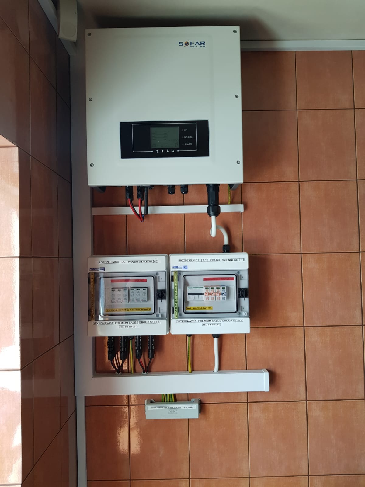 Zamontowany inwerter Sofar Solar z rozdzielnicami DC i AC na brązowej ścianie z płytek, widoczne przewody i elementy instalacji elektrycznej.