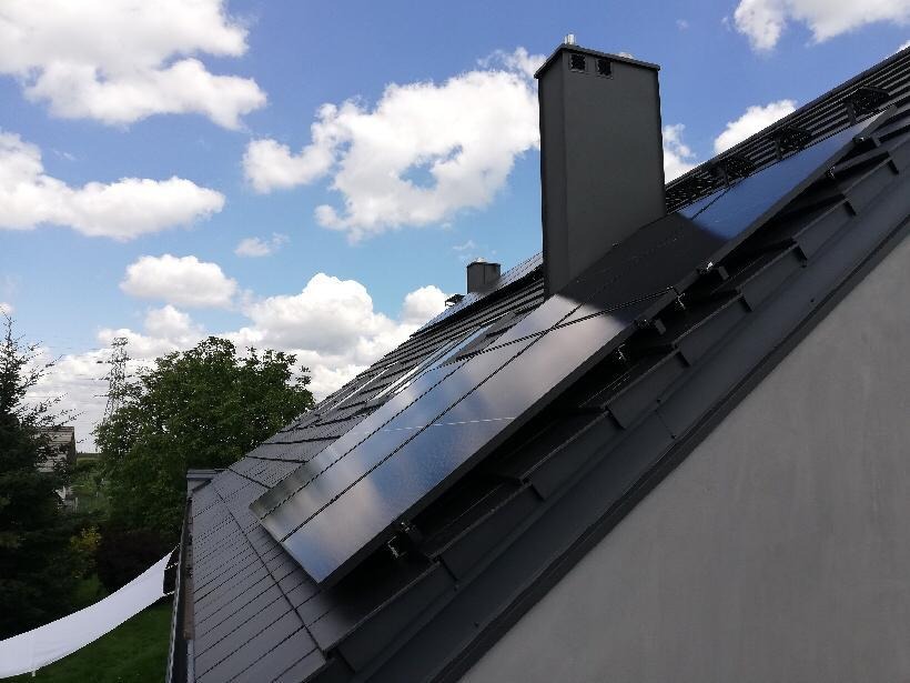 Panele słoneczne na ciemnym dachu domu jednorodzinnego, widoczne kominy i błękitne niebo z chmurami w tle.