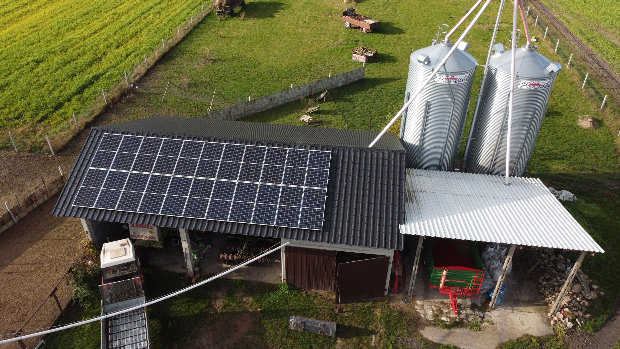 Widok z góry na budynek gospodarczy z panelami słonecznymi na dachu, obok silosy zbożowe, traktor i maszyny rolnicze w tle zielone pole.