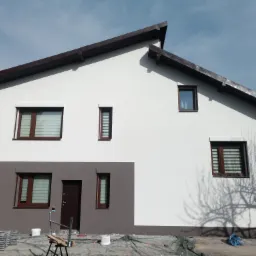 Dwukolorowa elewacja domu jednorodzinnego z brązowymi oknami i dachem, widoczne rusztowania i zabezpieczenia na ziemi, cień drzewa na ścianie.