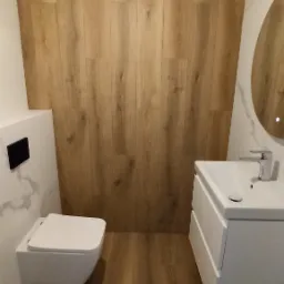 Nowoczesna łazienka z wiszącą toaletą, umywalką nablatową i okrągłym lustrem z podświetleniem, wykończona płytkami imitującymi drewno na ścianie i podłodze.