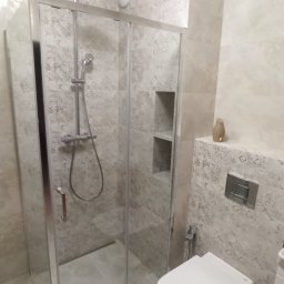 PRO-FLIZ - Szklana kabina prysznicowa z deszczownicą, wisząca toaleta i dekoracyjne płytki w odcieniach beżu i szarości w nowoczesnej łazience.