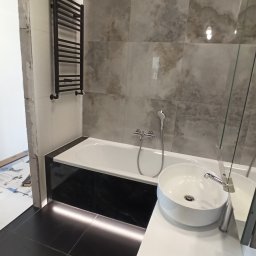 PRO-FLIZ - Nowoczesna łazienka z czarną armaturą, płytkami imitującymi marmur i podświetleniem LED pod wanną, widoczny fragment niezakończonego remontu.