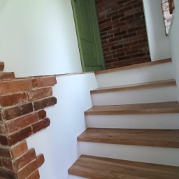 PRO-FLIZ - Schody z drewnianymi stopniami i białą podstopnicą, fragment ściany wykończony czerwoną cegłą, widoczna część zielonych drzwi i ściana z cegły w tle.