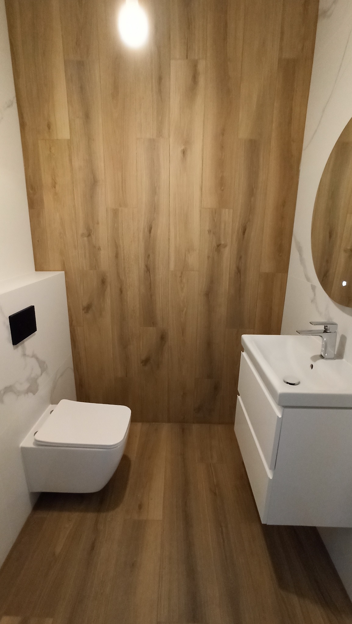 Nowoczesna łazienka z wiszącą toaletą, umywalką nablatową i okrągłym lustrem z podświetleniem, wykończona płytkami imitującymi drewno na ścianie i podłodze.