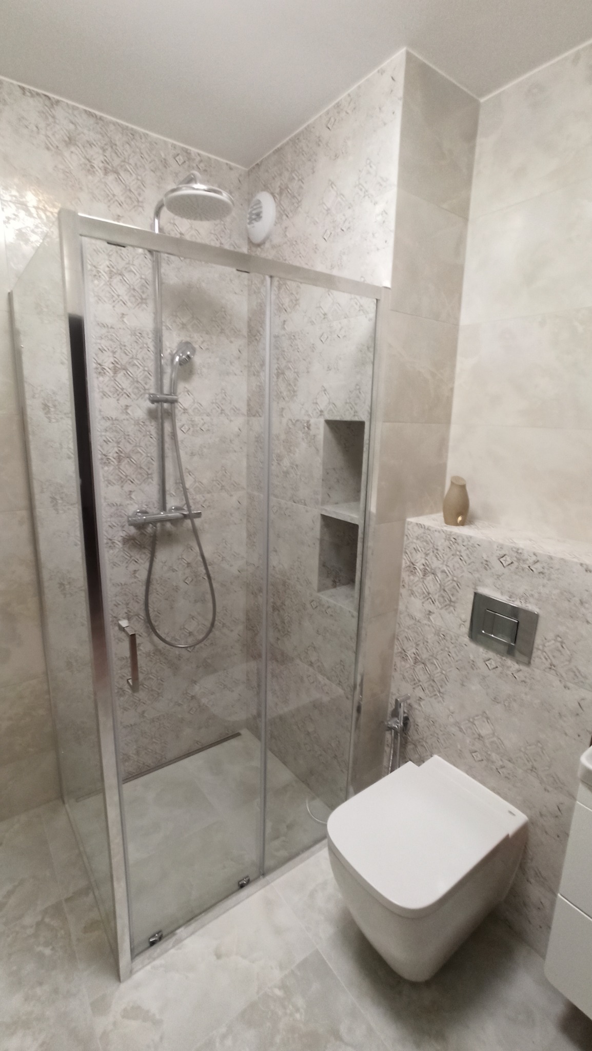 Szklana kabina prysznicowa z deszczownicą, wisząca toaleta i dekoracyjne płytki w odcieniach beżu i szarości w nowoczesnej łazience.