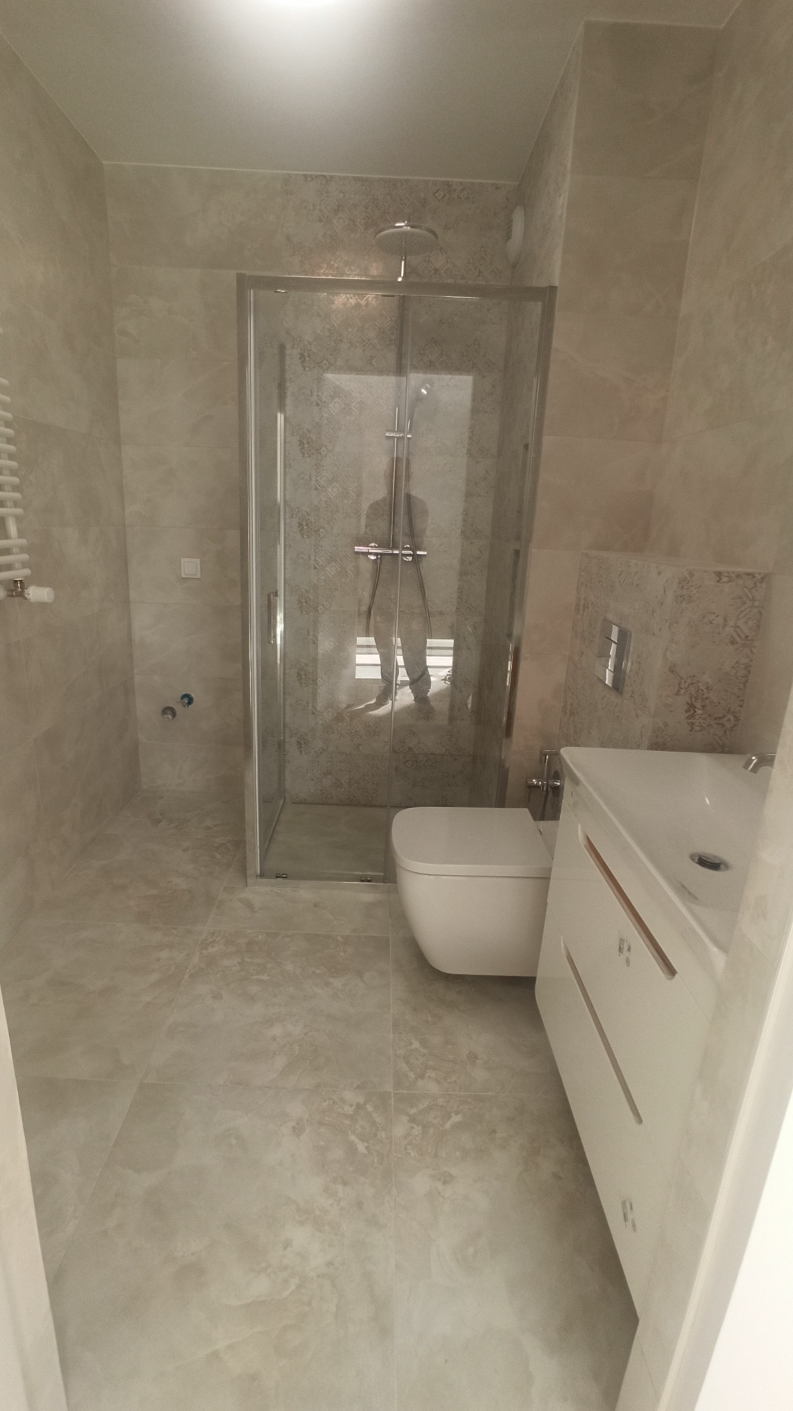 Wykończona łazienka w beżowych odcieniach z kabiną prysznicową ze szklanymi drzwiami, podwieszaną toaletą i umywalką z szafką, widoczne płytki imitujące kamień na ścianach i podłodze.