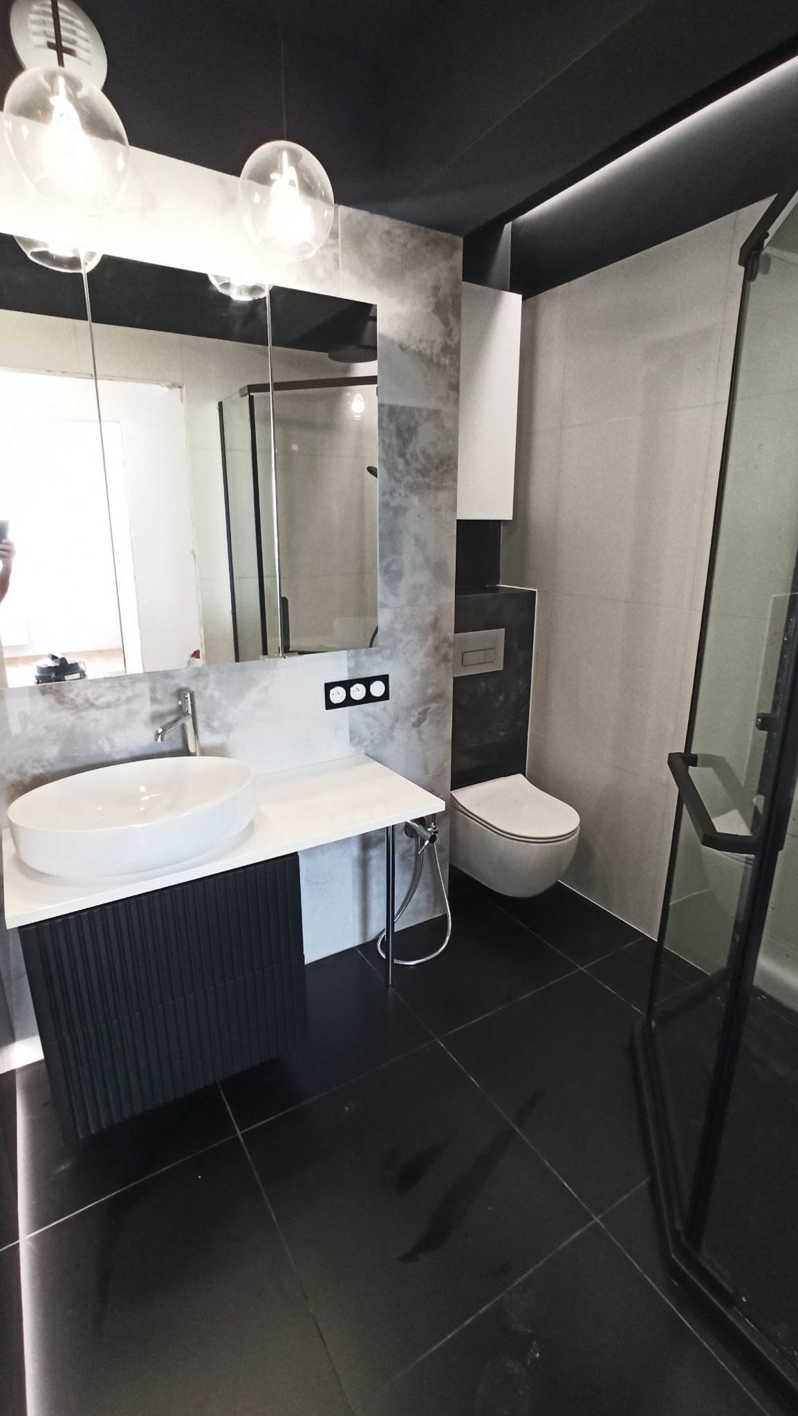 Nowoczesna łazienka z czarnymi płytkami podłogowymi, szarą mozaiką na ścianach, wiszącą białą miską WC, umywalką nablatową na czarnej szafce z ryflowanym frontem i szklaną kabiną prysznicową...