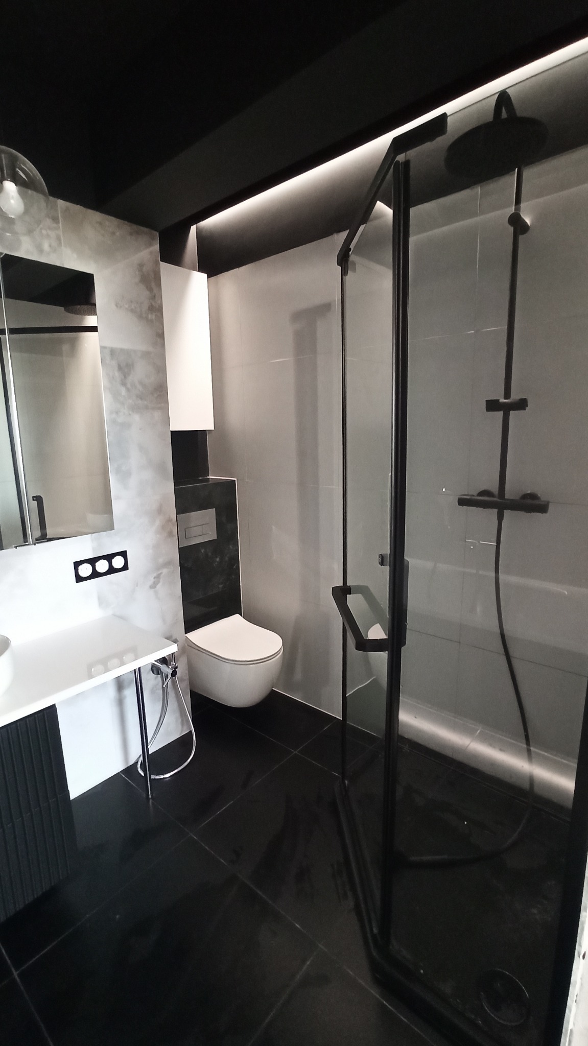 Nowoczesna łazienka z czarną armaturą, szarymi płytkami imitującymi marmur, czarną podłogą z połyskiem i kabiną prysznicową typu walk-in z czarnymi profilami.