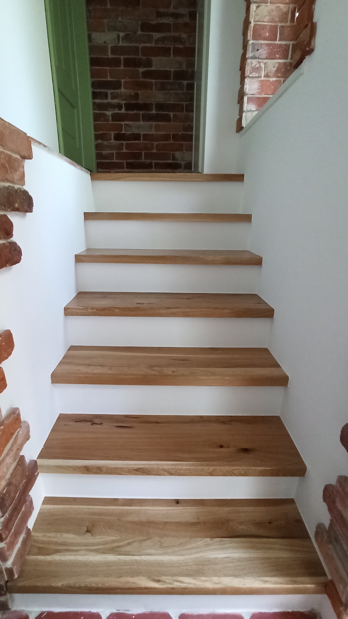 Schody z drewnianymi stopniami, białe ściany, fragment ceglanej ściany i zielone drzwi w tle.