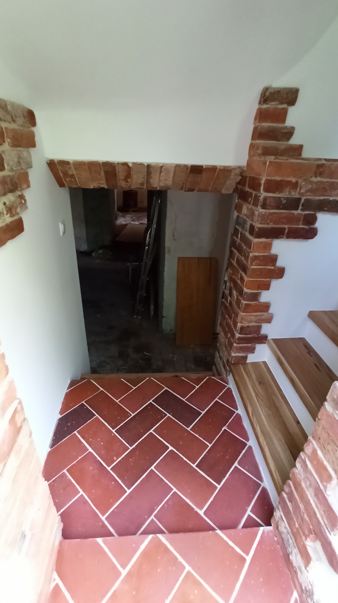 Ceglana podłoga ułożona w jodełkę, z fragmentem schodów wykończonych drewnem, ceglane obramowanie przejścia do innego pomieszczenia, w którym widać drabinę i płytę