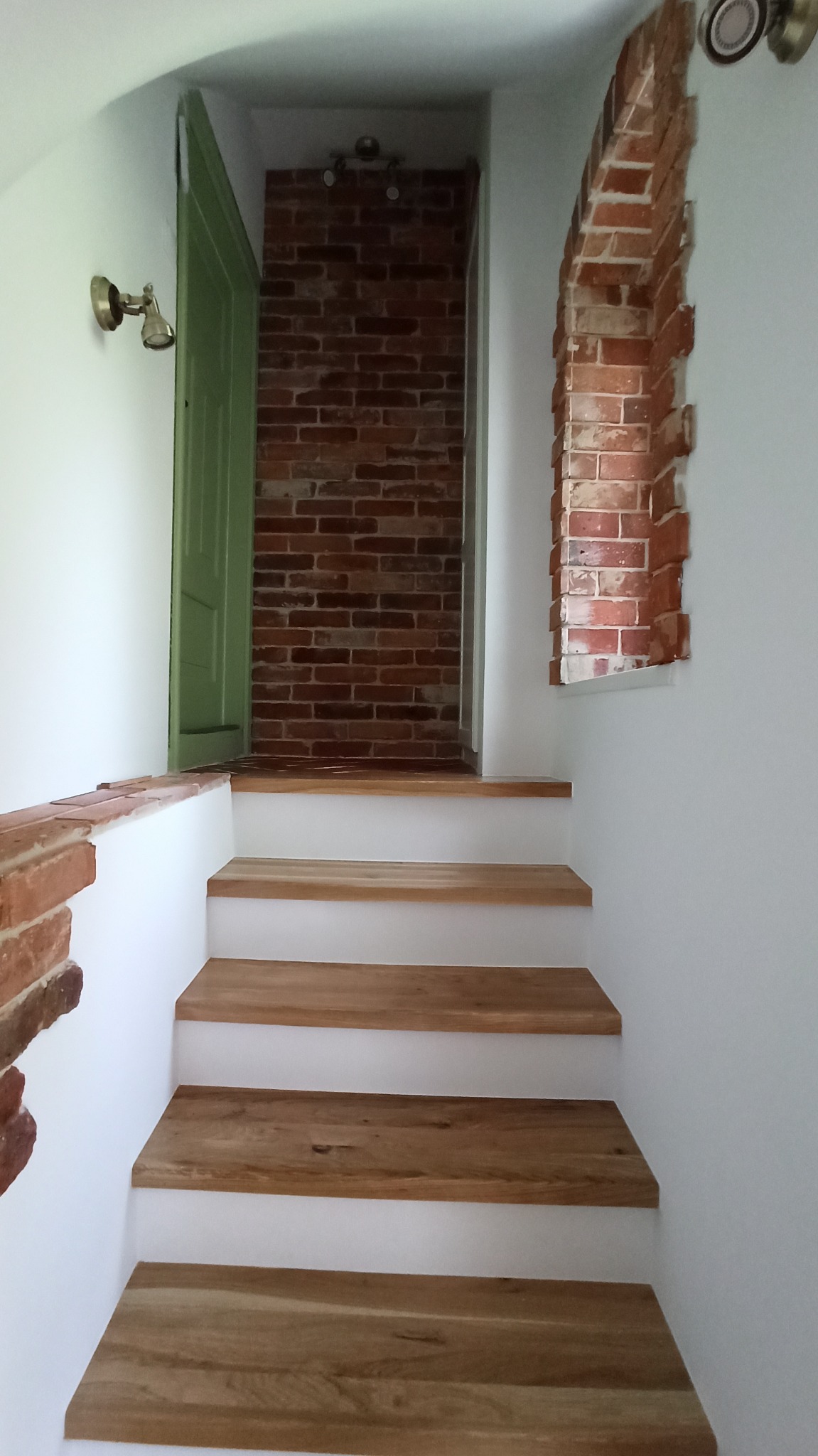 Schody drewniane z białymi podstopnicami prowadzące do łazienki z ceglaną ścianą i zielonymi drzwiami, obok ceglany łuk w ścianie.