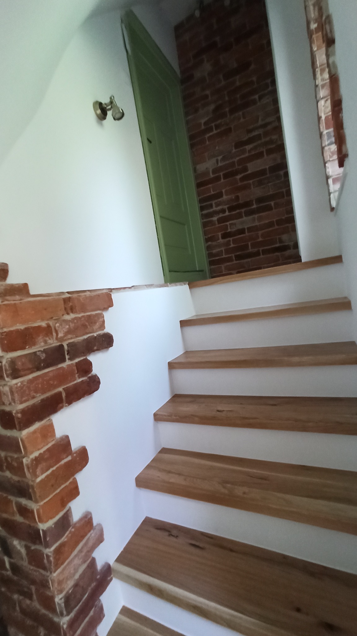 Schody z drewnianymi stopniami i białą podstopnicą, fragment ściany wykończony czerwoną cegłą, widoczna część zielonych drzwi i ściana z cegły w tle.