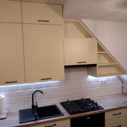 Usługi Instalacyjno-Remontowe Wojciech Struski - Jasna kuchnia na poddaszu z białymi płytkami metra, czarnym zlewem i płytą gazową, oświetleniem LED pod szafkami i skośnymi półkami.