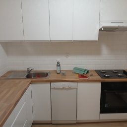 Usługi Instalacyjno-Remontowe Wojciech Struski - Wnętrze kuchni po remoncie: białe szafki, drewniany blat, białe płytki na ścianie, zamontowana płyta gazowa i piekarnik, widoczne ślady po pracy, butelka ze środkiem czyszczącym, głośnik JBL...