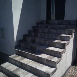 Usługi Instalacyjno-Remontowe Wojciech Struski - Zewnętrzne schody wejściowe wyłożone płytkami w odcieniach szarości, prowadzące do ciemnych drzwi wejściowych, oświetlone naturalnym światłem z wyraźnym cieniem na ścianie.