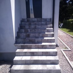 Usługi Instalacyjno-Remontowe Wojciech Struski - Wyłożone szarymi płytkami schody wejściowe do domu z ciemnymi drzwiami, widok z dołu, obok ścieżka z kamyków.