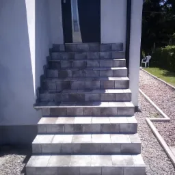 Wyłożone szarymi płytkami schody wejściowe do domu z ciemnymi drzwiami, widok z dołu, obok ścieżka z kamyk&oacute;w.