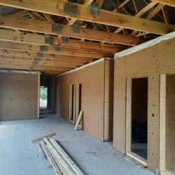 Usługi Instalacyjno-Remontowe Wojciech Struski - Wnętrze domu w trakcie budowy, widoczne drewniane elementy konstrukcyjne dachu oraz ściany wyłożone płytami z materiału izolacyjnego o brązowej strukturze, przygotowane otwory na drzwi i okna...