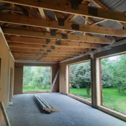 Wnętrze w trakcie ocieplania poddasza, widoczne drewniane krokwie z metalowymi łącznikami, ściany wyłożone płytami izolacyjnymi, duże okna z widokiem na zieleń, drabina oparta o ścianę, deski...