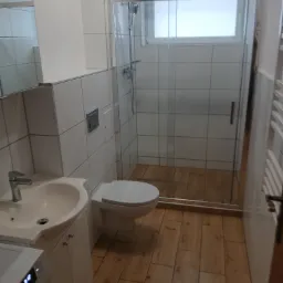 Wykończona łazienka z kabiną prysznicową, umywalką z szafką, toaletą i pralką, wyłożona białymi płytkami i drewnopodobną podłogą.