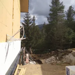 Ocieplanie ściany budynku styropianem z widocznymi elementami konstrukcyjnymi, rurami i deskami na placu budowy w tle z drzewami i pochmurnym niebem.