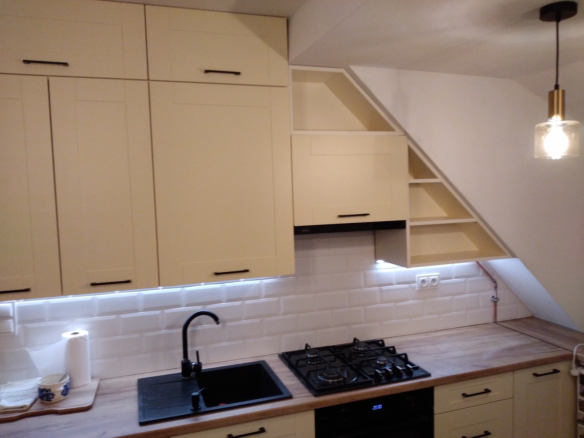 Jasna kuchnia na poddaszu z białymi płytkami metra, czarnym zlewem i płytą gazową, oświetleniem LED pod szafkami i skośnymi półkami.