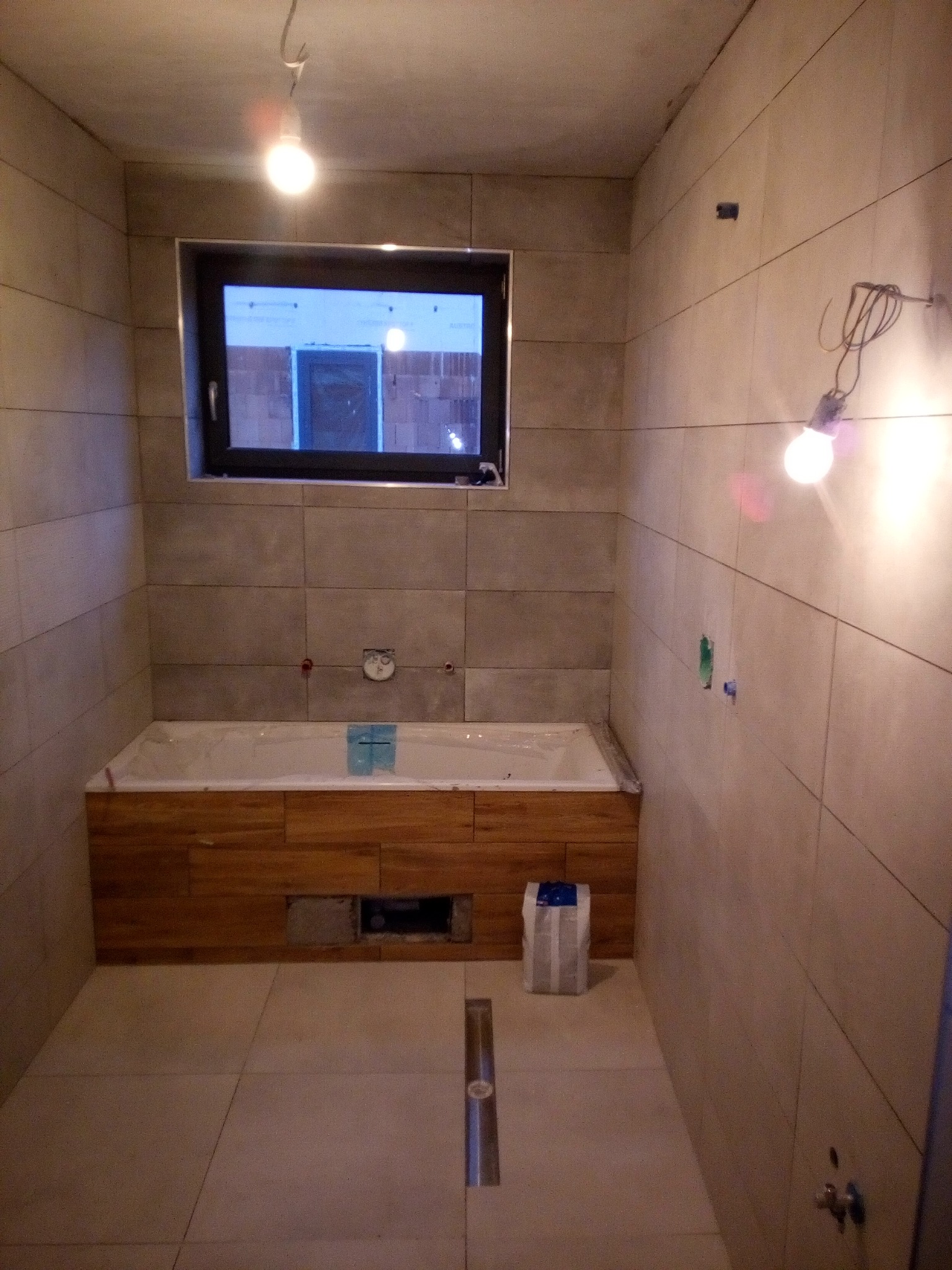 Łazienka w trakcie prac wykończeniowych: ułożone płytki ścienne i podłogowe w odcieniach beżu, obudowana drewnem wanna, widoczne wystające kable i żarówka, worek z zaprawą na podłodze.