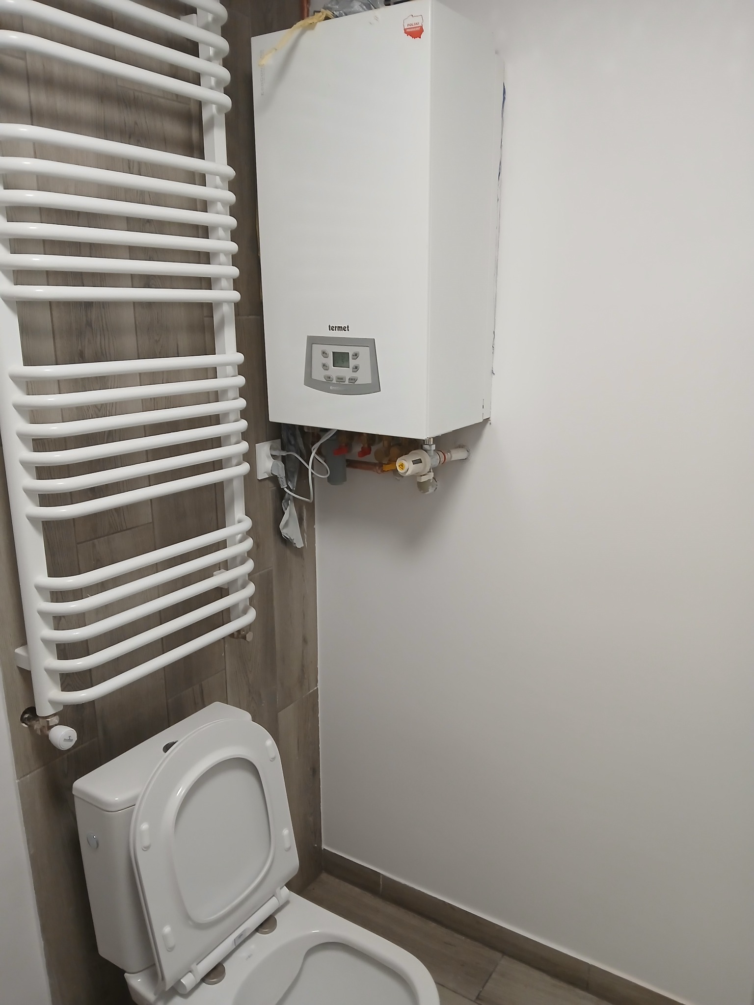Biały piec gazowy Termet zamontowany na ścianie nad toaletą w łazience, obok grzejnik drabinkowy, widoczne przyłącza wodne i gazowe, płytki imitujące drewno na fragmencie ściany.