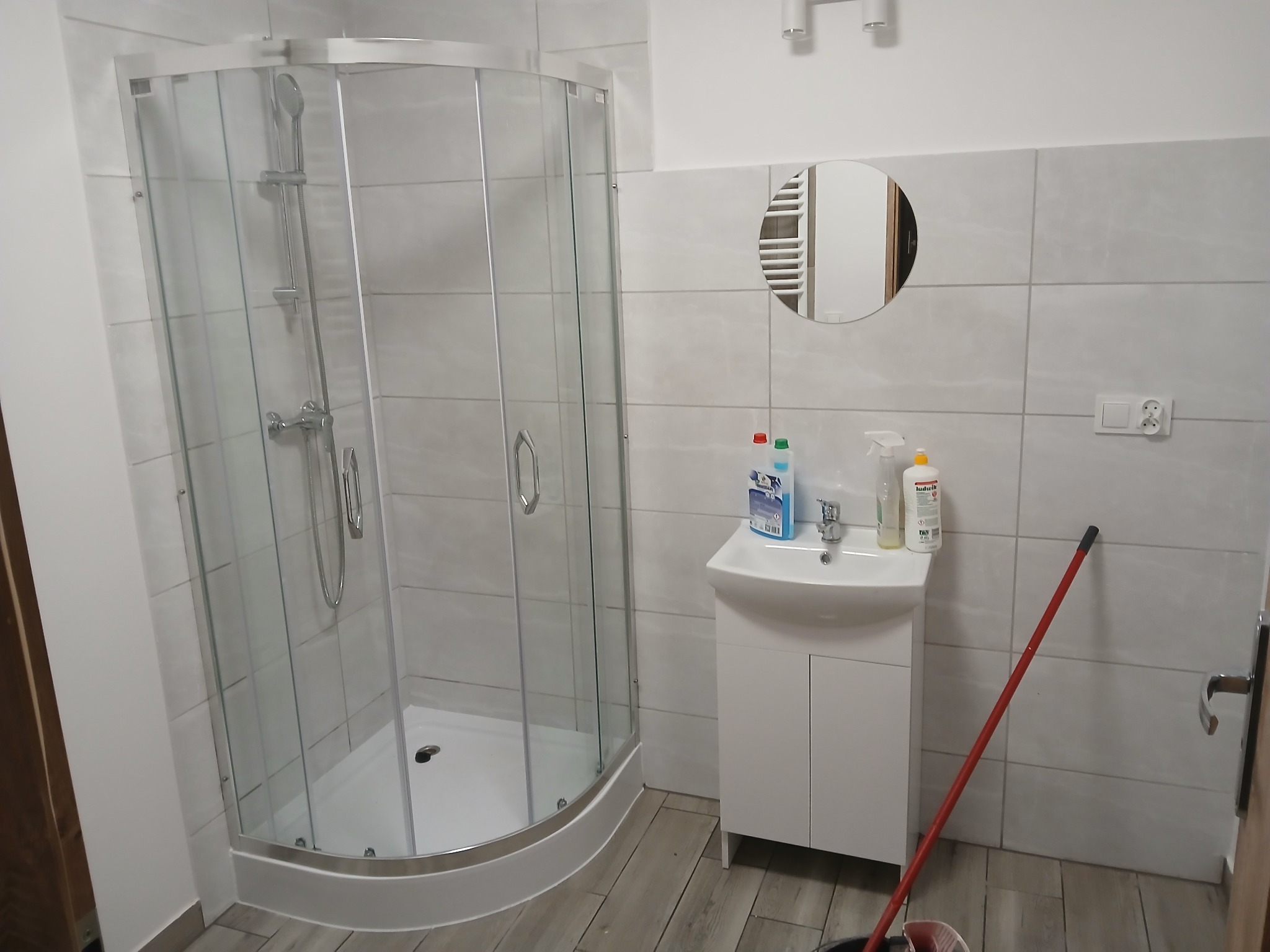 Nowoczesna łazienka z kabiną prysznicową z zaokrąglonym szkłem, białą umywalką z szafką, okrągłym lustrem i szarymi płytkami na ścianach i podłodze, widoczna mop oparty o ścianę.