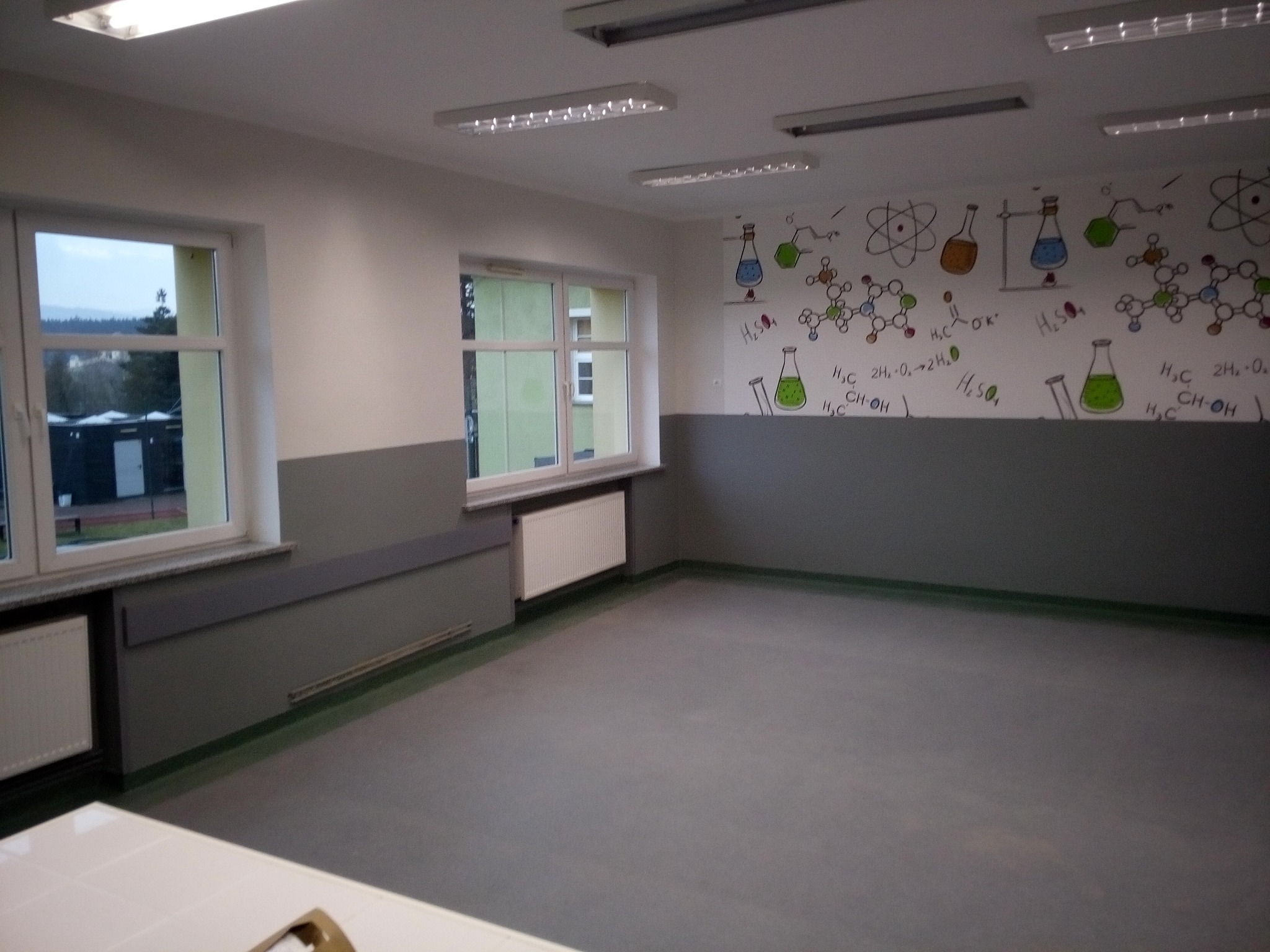 Pomalowane ściany w sali lekcyjnej z motywami chemicznymi, widoczne okna i grzejniki, szare panele ścienne, jasne oświetlenie sufitowe.