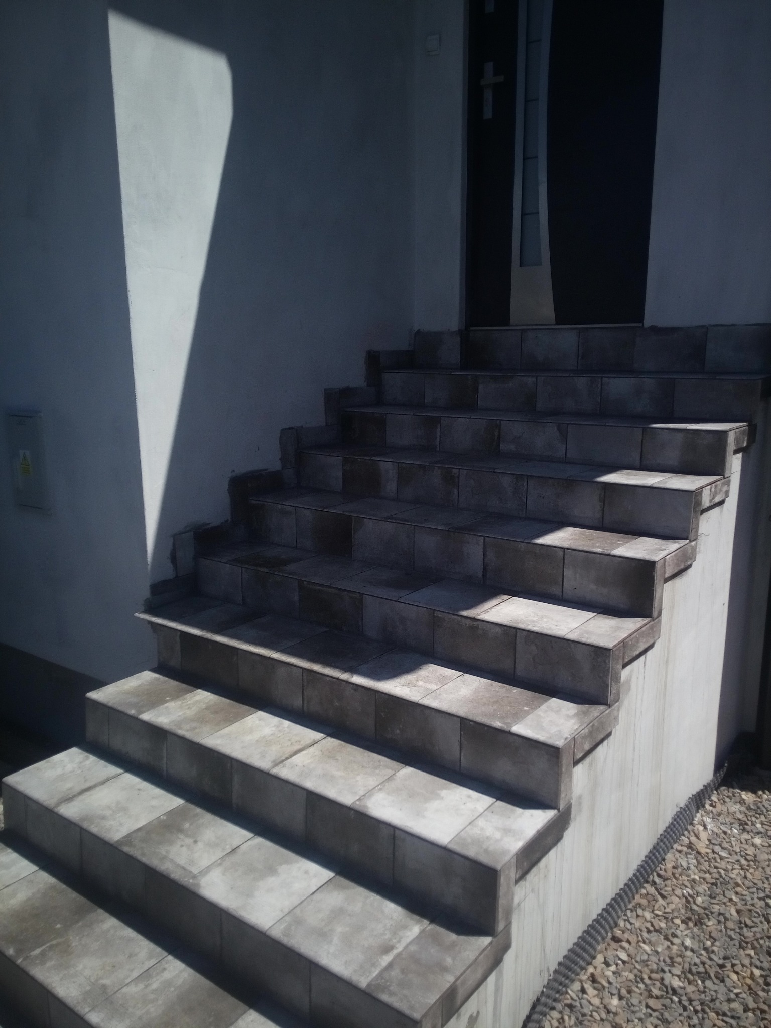 Zewnętrzne schody wejściowe wyłożone płytkami w odcieniach szarości, prowadzące do ciemnych drzwi wejściowych, oświetlone naturalnym światłem z wyraźnym cieniem na ścianie.