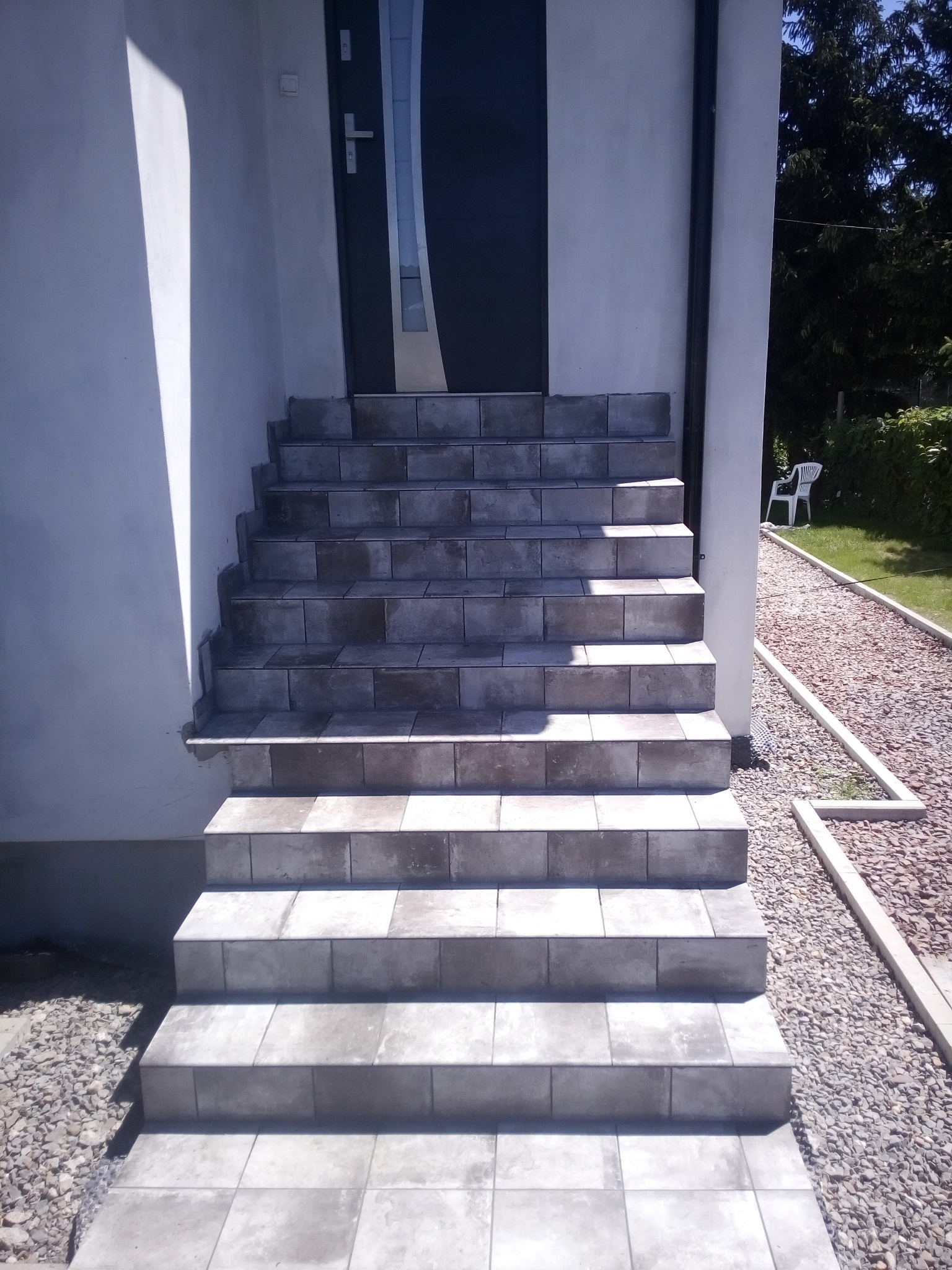Wyłożone szarymi płytkami schody wejściowe do domu z ciemnymi drzwiami, widok z dołu, obok ścieżka z kamyków.