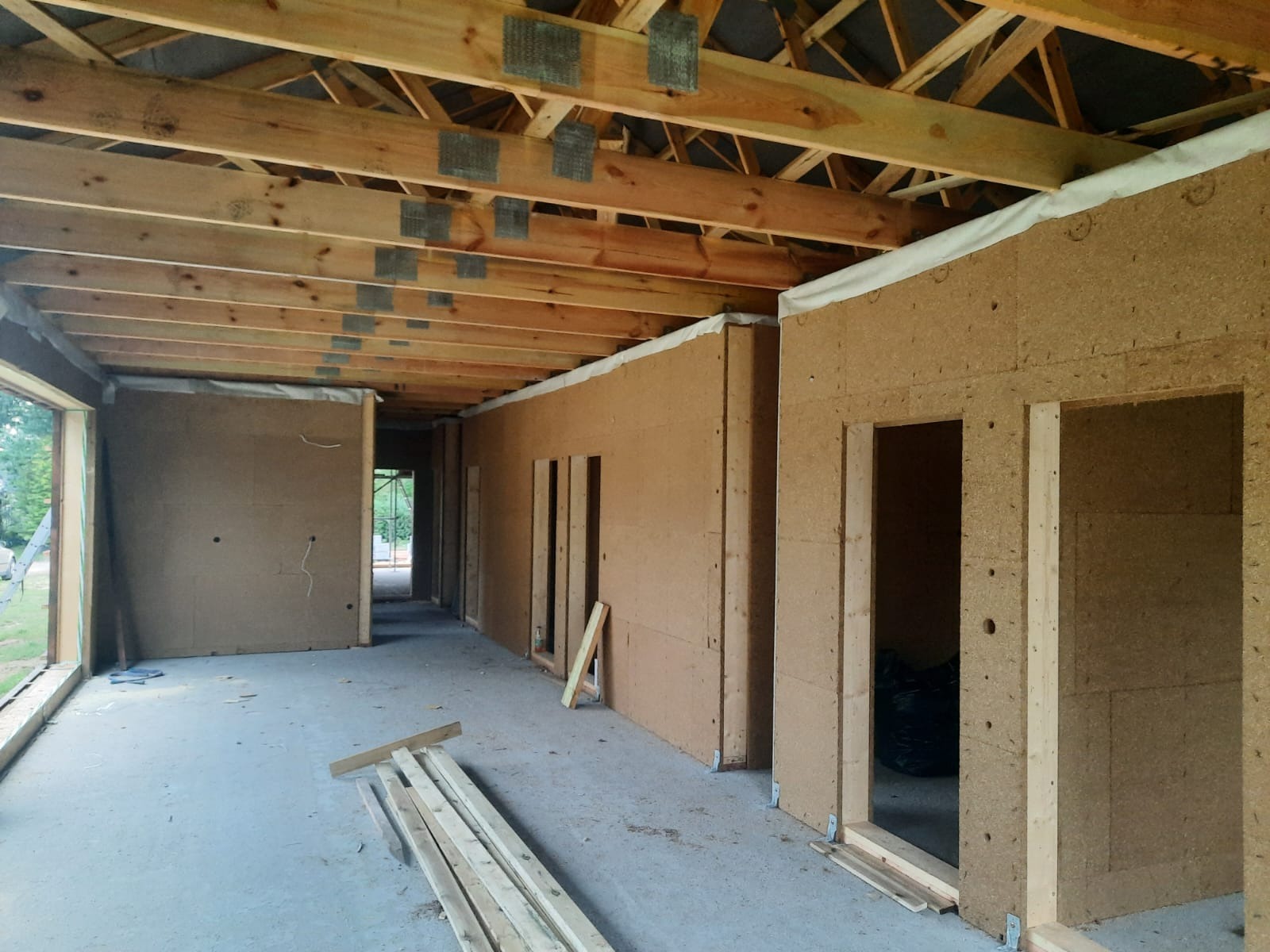 Wnętrze domu w trakcie budowy, widoczne drewniane elementy konstrukcyjne dachu oraz ściany wyłożone płytami z materiału izolacyjnego o brązowej strukturze, przygotowane otwory na drzwi i okna...