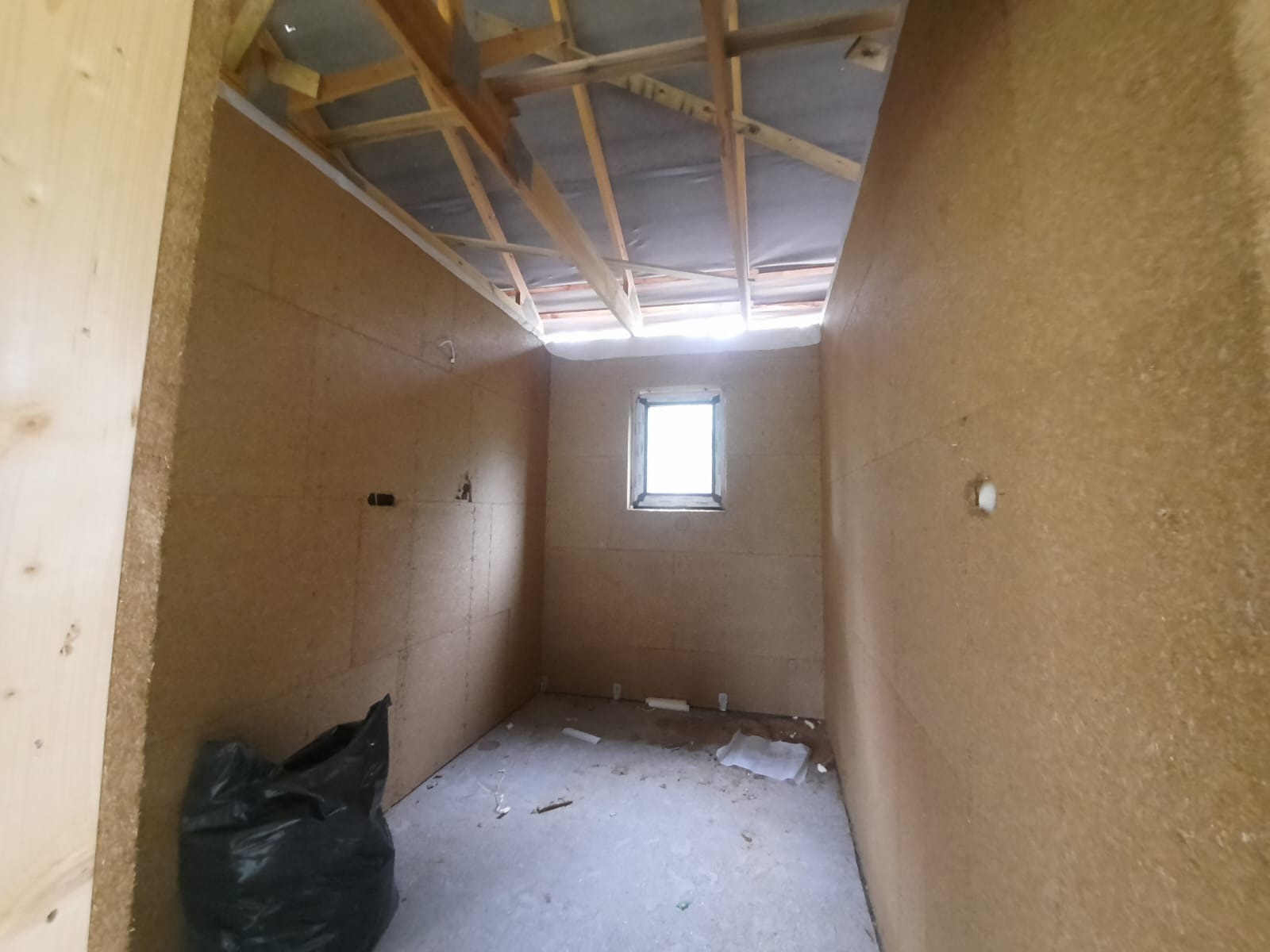 Poddasze w trakcie ocieplania, widoczne płyty izolacyjne na ścianach, surowy strop z drewnianą konstrukcją i folią, okno w ścianie szczytowej, czarny worek z odpadami w rogu pomieszczenia.