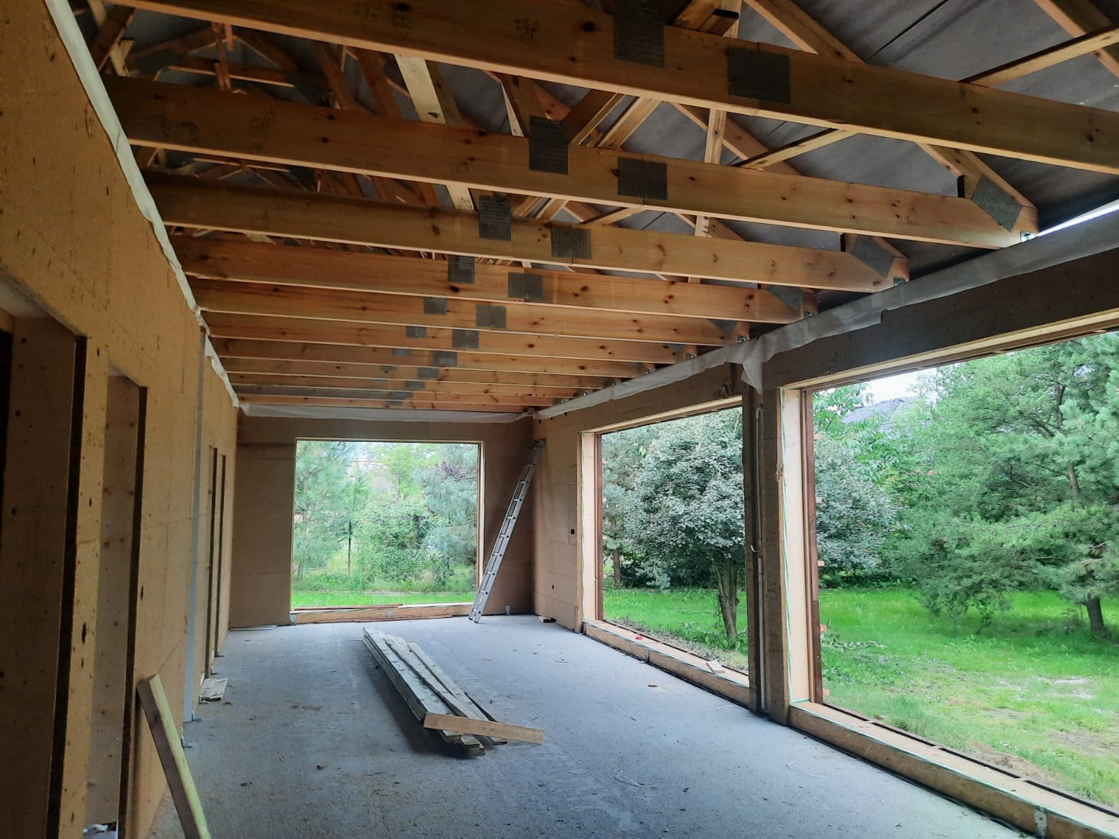 Wnętrze w trakcie ocieplania poddasza, widoczne drewniane krokwie z metalowymi łącznikami, ściany wyłożone płytami izolacyjnymi, duże okna z widokiem na zieleń, drabina oparta o ścianę, deski...