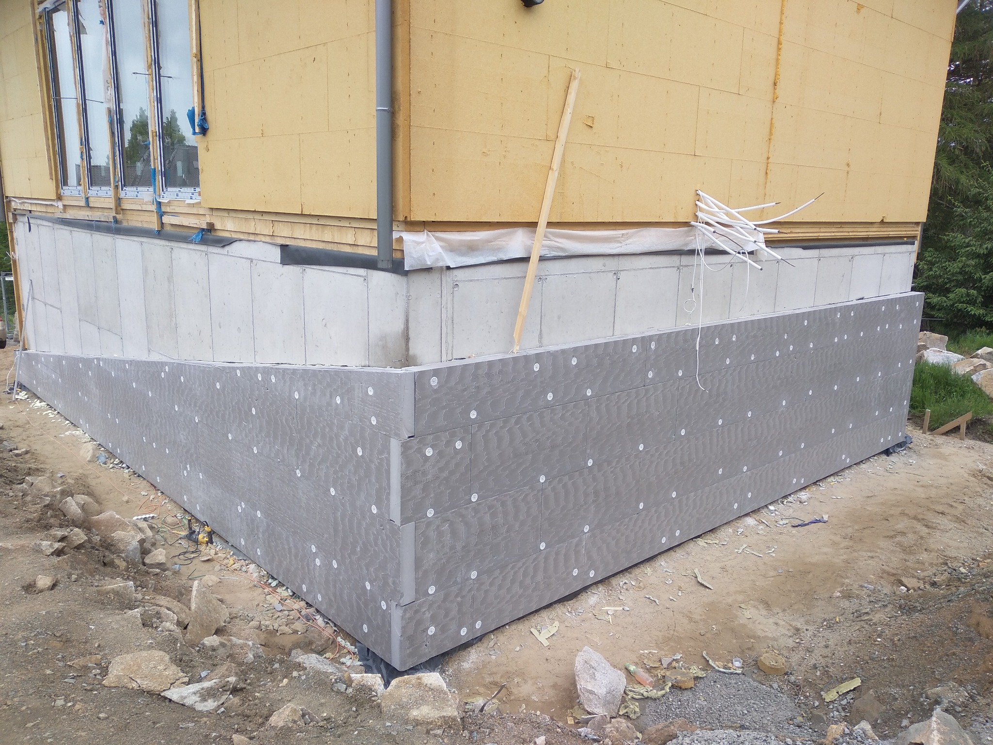 Ocieplenie fundamentu budynku płytami izolacyjnymi z widocznymi kołkami mocującymi, teren wokół w stanie surowym.